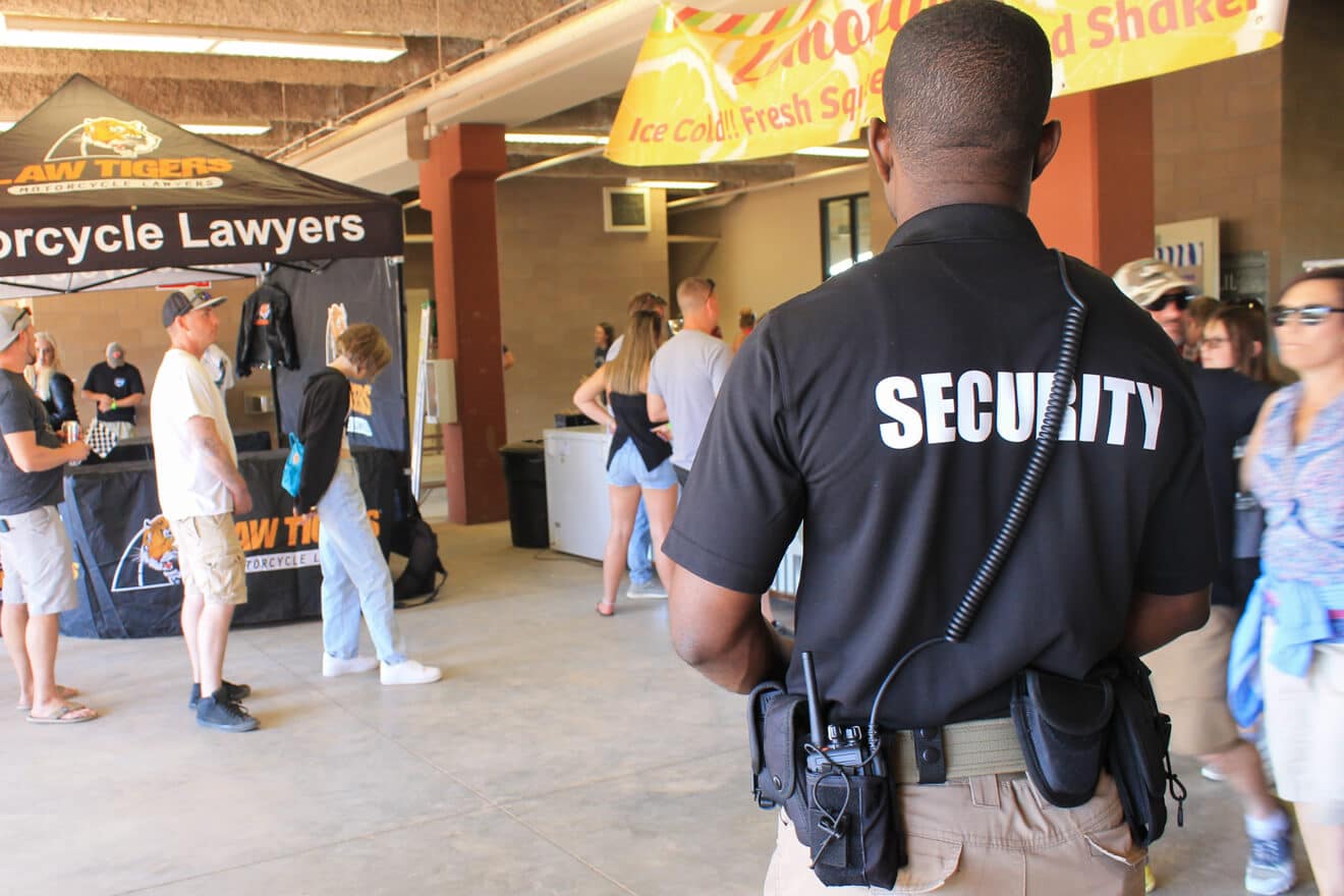 A man with a shirt that says "security" across the back is standing at the entrance of an event and watching all the attendees entering the event.