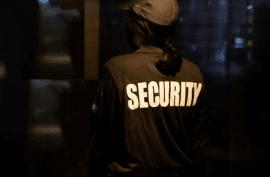 The image shows the back of a security guard wearing a black jacket and a hat and on the jacket in large, white letters is the word "Security".