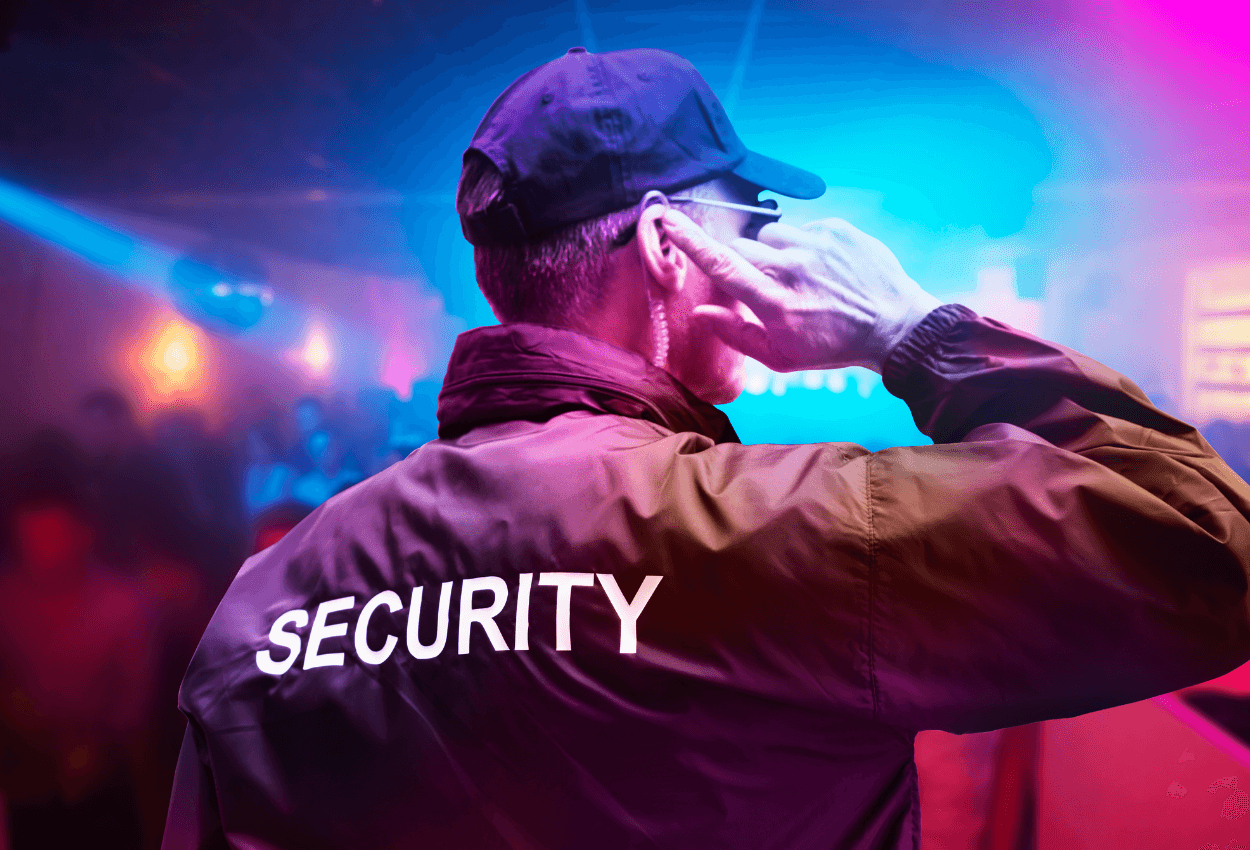 The image shows the back of a male bar security guard. He's wearing a black jacket and the word "security" is written across the back in white. There are colorful lights from the bar in the background.