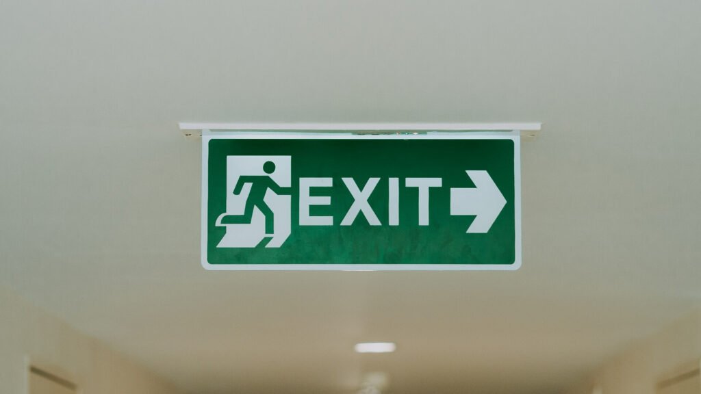 A green "exit" sign on a hallway ceiling. The sign has a cartoon man running through a door.