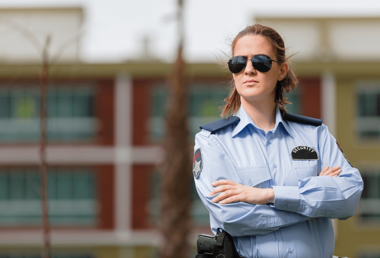 The image shows a workplace security guard standing outside with her arms crossed and she has sunglasses on.