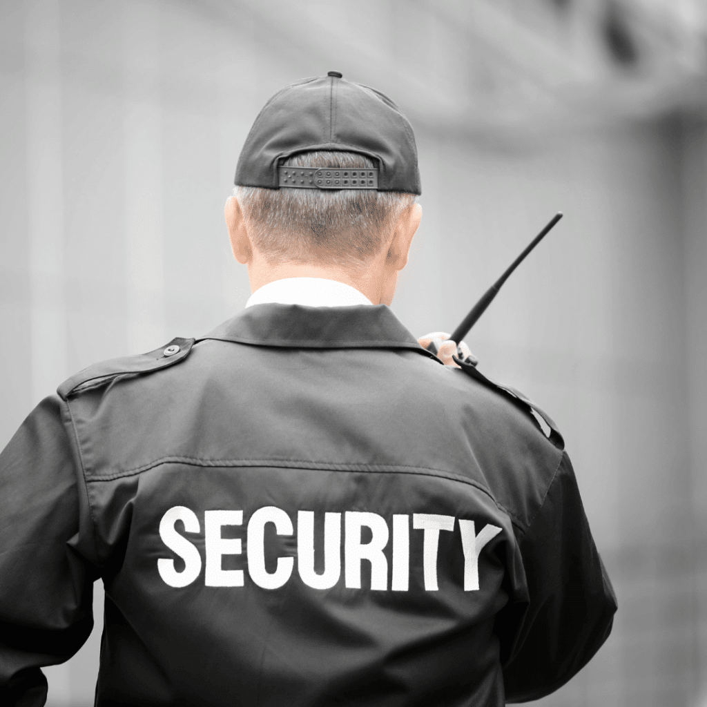 A photo of a man from the back using a walkie talkie and wearing a black jacket that says "Security" in white letters
