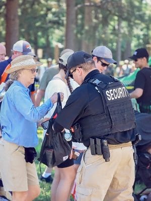 A security officer checks people's belongings in a wooded area, at a security checkpoint with a green van in the background.