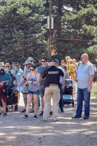 Security officer conducting bag checks at an outdoor event entrance as a diverse group of attendees wait in line under sunny weather.