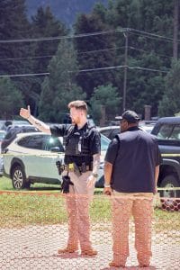 Security guard directing event attendees near a temporary orange fence, with another staff member beside him and parked vehicles in the background.