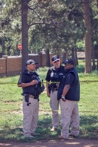 Three security personnel in uniform standing together outdoors, having a discussion near caution tape in a shaded grassy area.