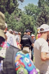 Security officer monitoring a crowded outdoor event, surrounded by attendees in casual clothing under a canopy of green trees.