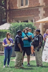 Security personnel talking with event attendees on a grassy area near a historic brick building, with volunteers wearing colorful t-shirts and holding food containers.