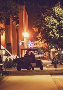 Two security personnel riding a golf cart along a dimly lit city sidewalk at night, with warm streetlights and brick buildings in the background.