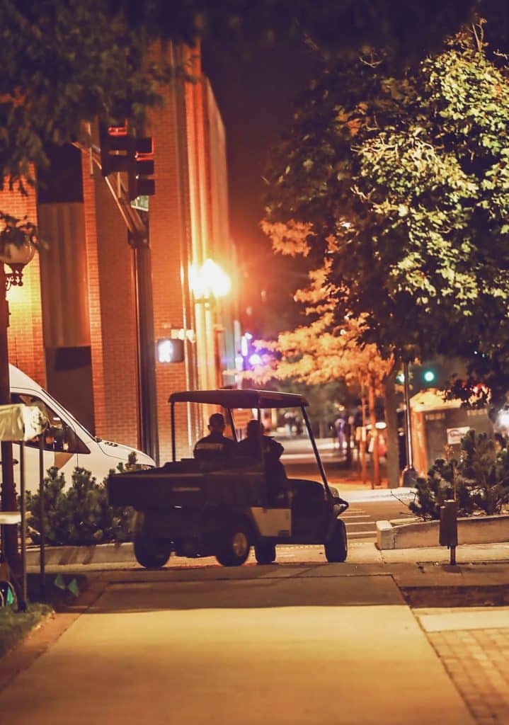 Two security personnel riding a golf cart along a dimly lit city sidewalk at night, with warm streetlights and brick buildings in the background.