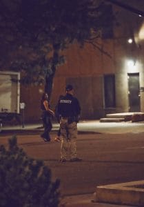 Security officer monitoring a quiet urban street at night while two women walk in the background near a dimly lit building entrance.