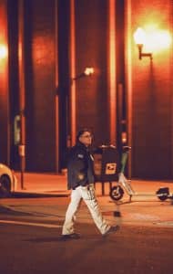 A security guard walking across a downtown street at night, illuminated by orange streetlights, with a scooter and mailbox in the background.