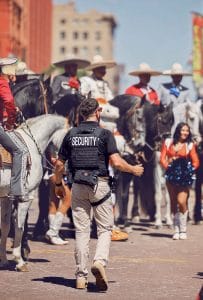 Security guard overseeing a vibrant street parade scene with horseback riders in traditional charro outfits and a cheerleader in front.
