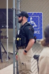 Bearded security officer standing in front of a chain-link fence near a handicap accessible sign, dressed in tactical gear with sunglasses and a cap.