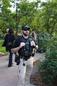 Security officer standing in a park area during an outdoor event, wearing tactical gear and sunglasses with string lights and trees in the background.
