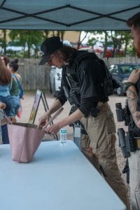 Security staff inspecting a pink handbag at a check-in table under a tent, as event attendees wait in the background.
