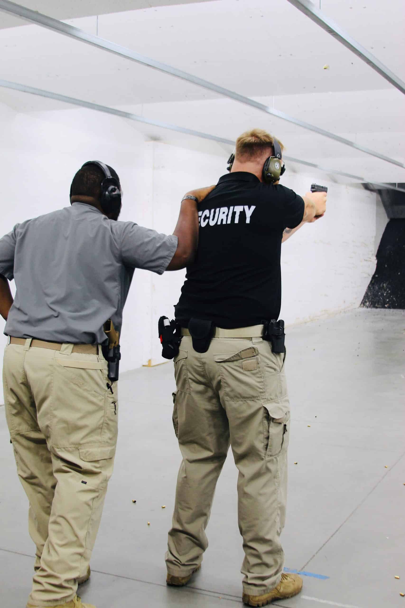 Two men in a shooting range, one aiming a handgun, both wearing protective gear.