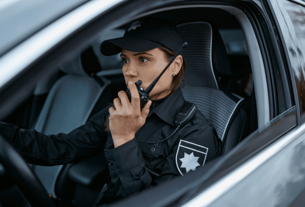 A picture of a security guard patrolling in her car as she talks into a walkie talkie.