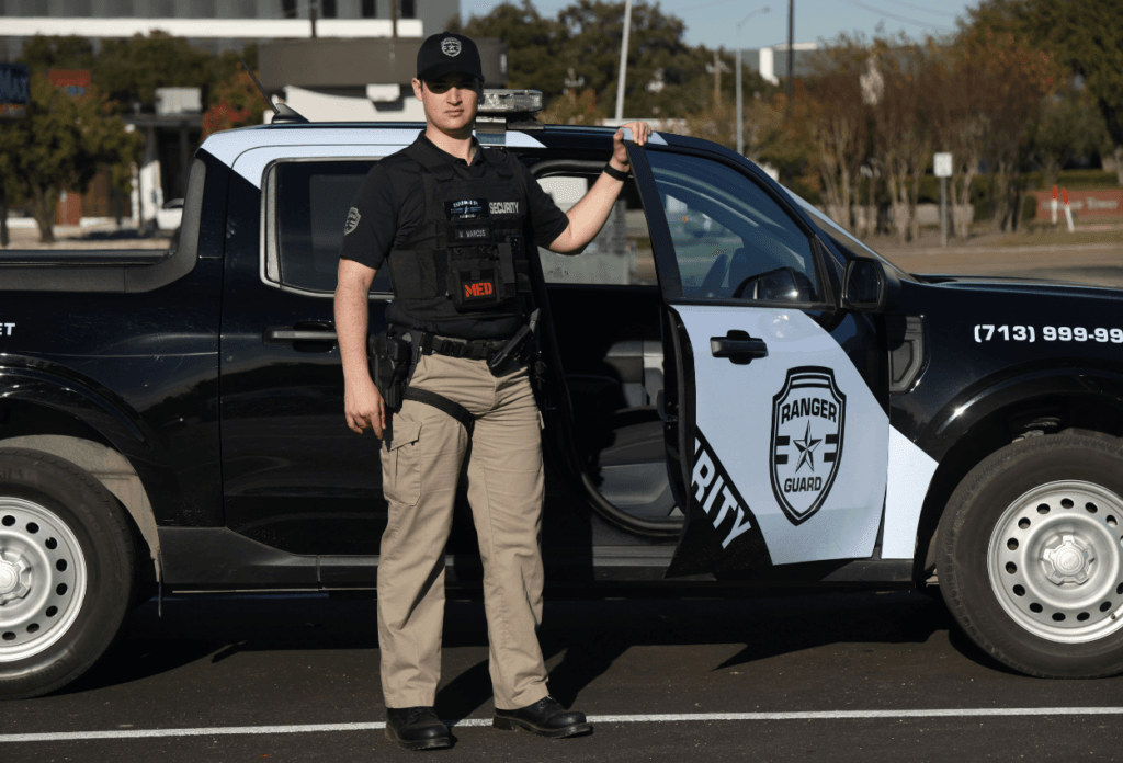 A picture of a security guard standing outside his car holding the door and looking into a camera.