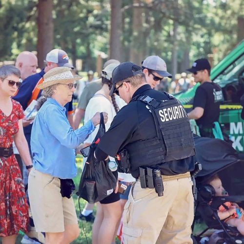 A security officer checks people's belongings in a wooded area, at a security checkpoint with a green van in the background.
